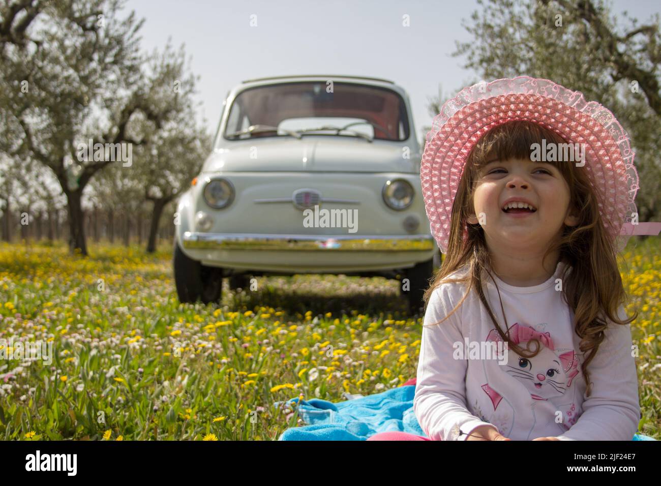 Adorable and beautiful smiling little girl sitting in a flowery meadow with an old Italian Fiat 500 car in the background. Vacation in Florence, Stock Photo