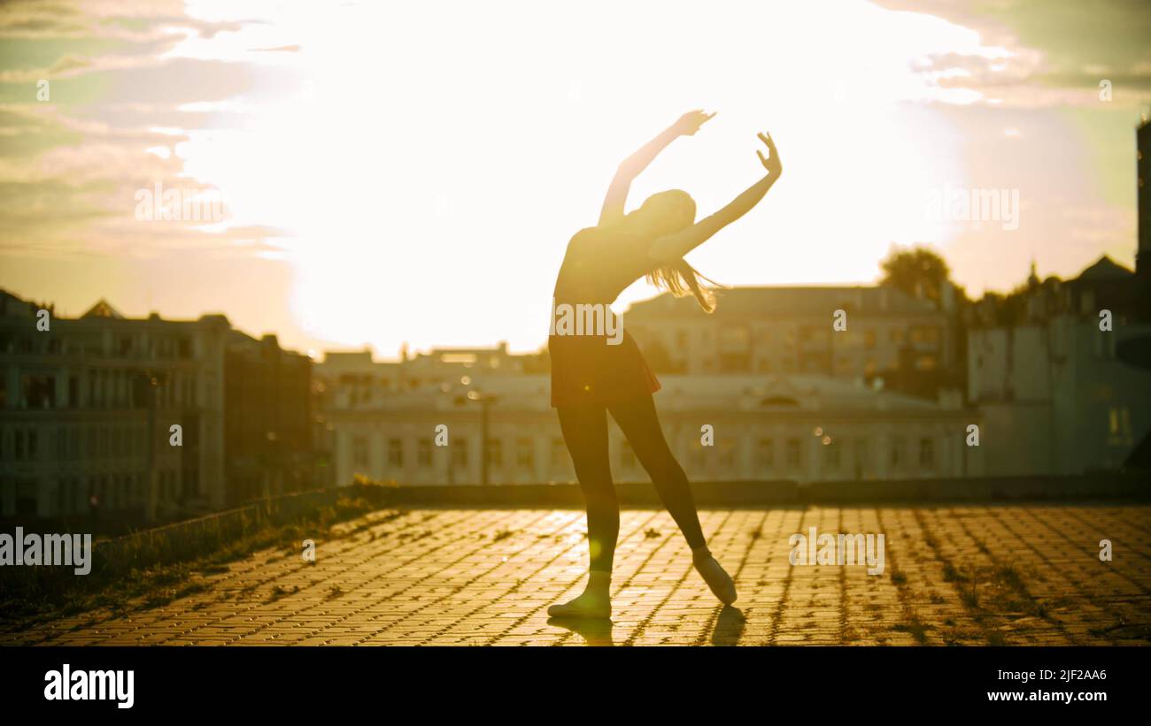 A silhouette of young woman ballerina in dress standing in the graceful pose on the roof on a background of modern buildings - bright sunset Stock Photo