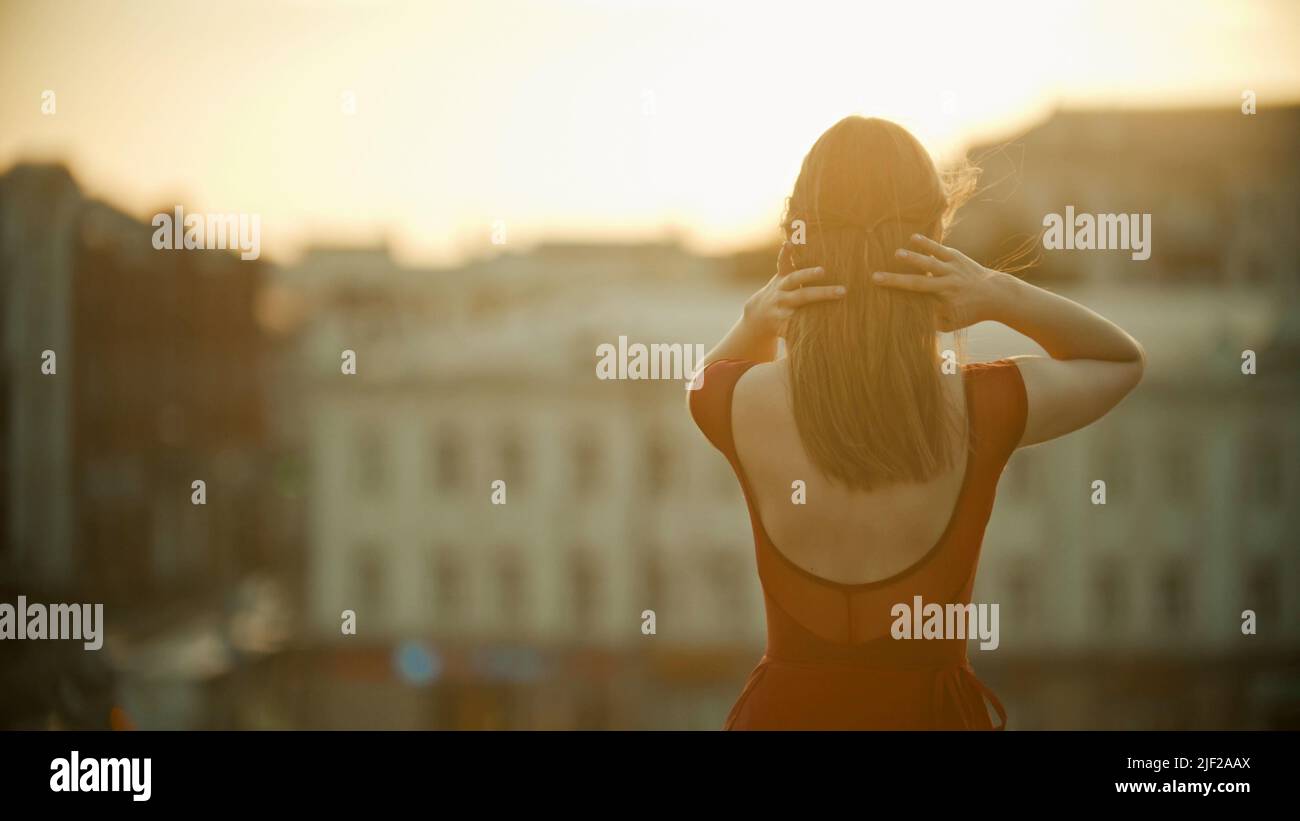 A young woman in red dress standing on the roof and fixing her hair - sunset Stock Photo