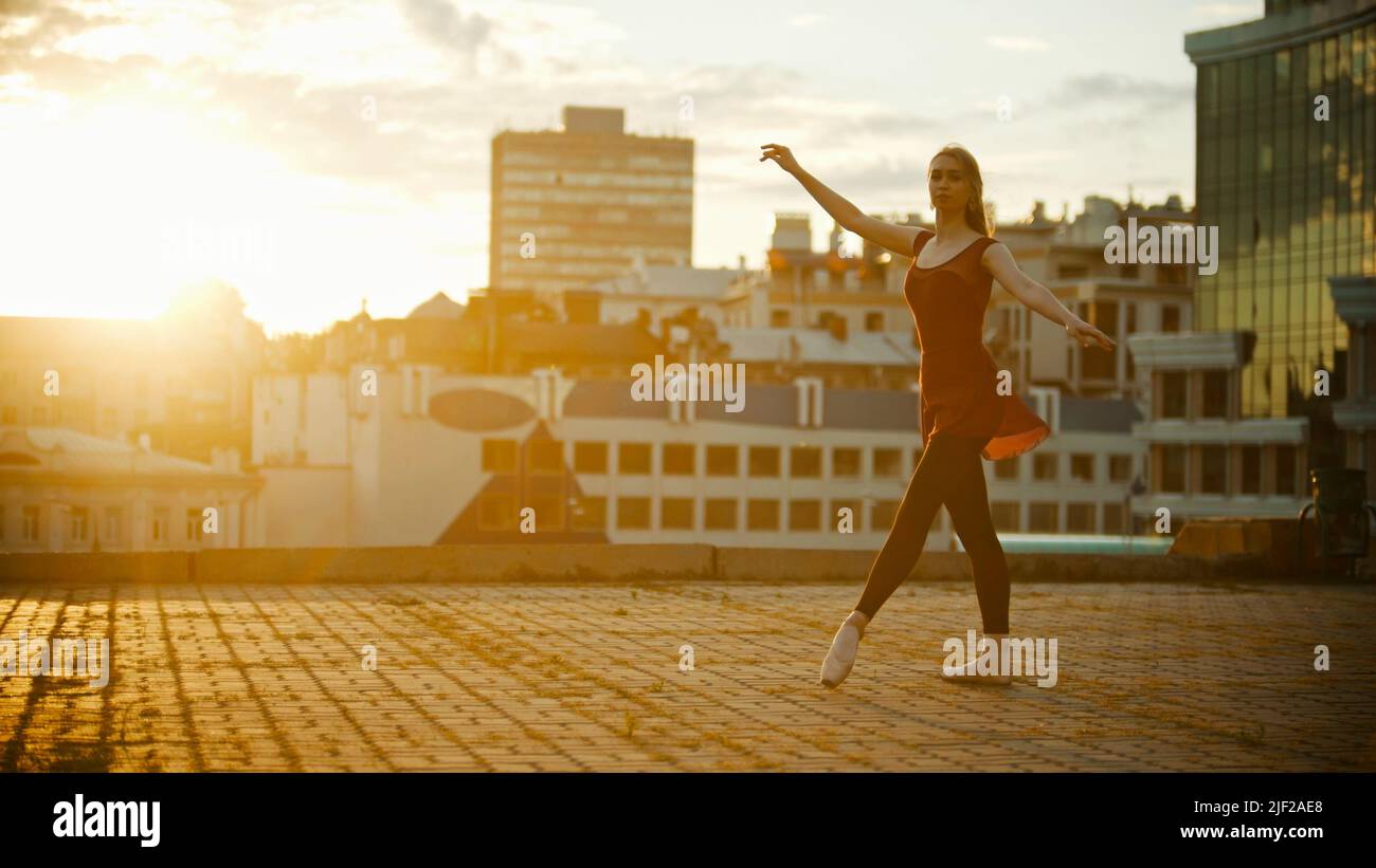 Young graceful woman in red dress ballerina standing in the pose on the roof - bright sunset Stock Photo