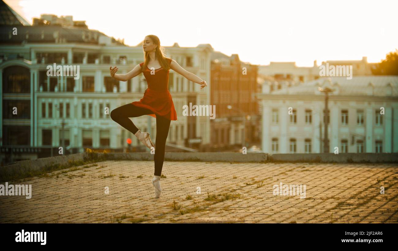 Young inspired woman in red dress ballerina standing in the graceful pose on the roof - modern buildings on the background - bright sunset Stock Photo