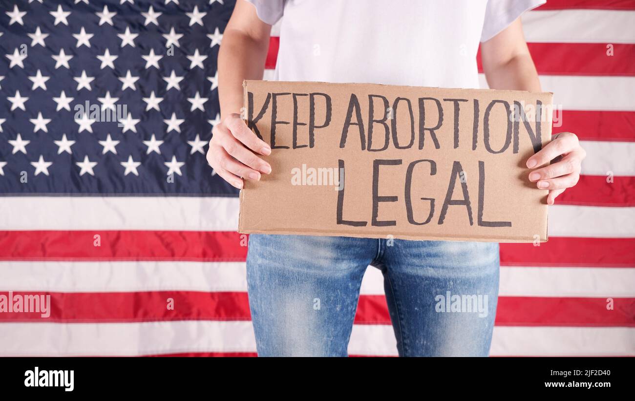 Young woman protester holds cardboard with Keep Abortion Legal sign against USA flag on background. Girl protesting against anti-abortion laws Stock Photo