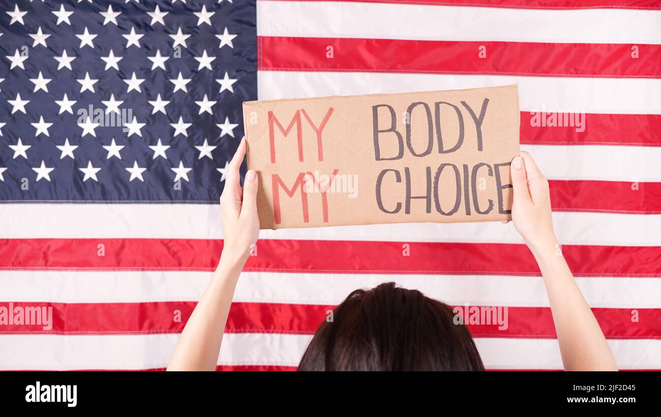 Young woman protester holds cardboard with My Body My Choice sign against USA flag on background. Girl protesting against anti-abortion laws. Feminist Stock Photo