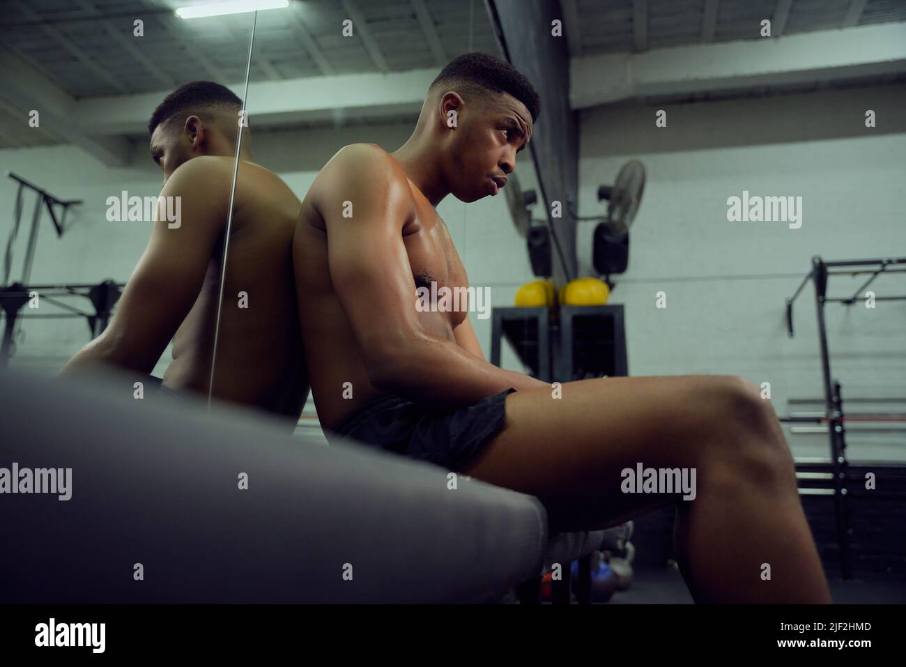 Young African American male athlete resting in the gym after a hard cross training. Shirtless mixed race male sitting down after exercising. High Stock Photo