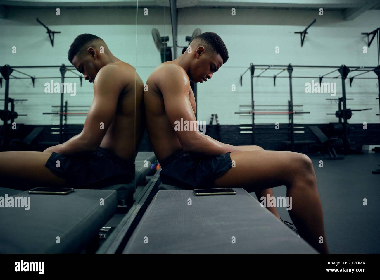 Young African American male athlete resting in the gym after a hard cross training. Shirtless mixed race male sitting down after exercising. High Stock Photo