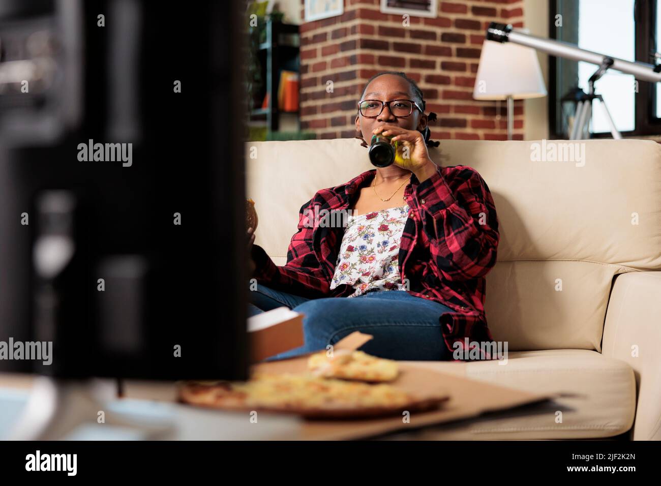 Cheerful adult drinking alcoholic beer from bottle on couch, watching movie  or film on television channel. Eating fast food meal from delivery package  and enjoying beverage in living room Stock Photo -