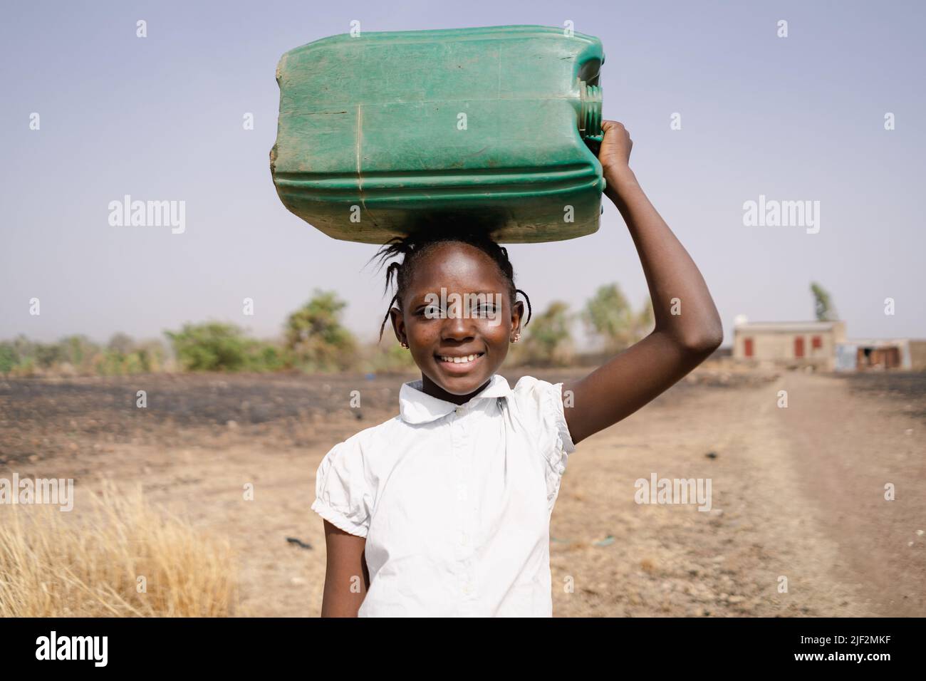 Smiling African village girl carrying a large green canister on her head as she walks to the community water station; human right to safe drinking wat Stock Photo
