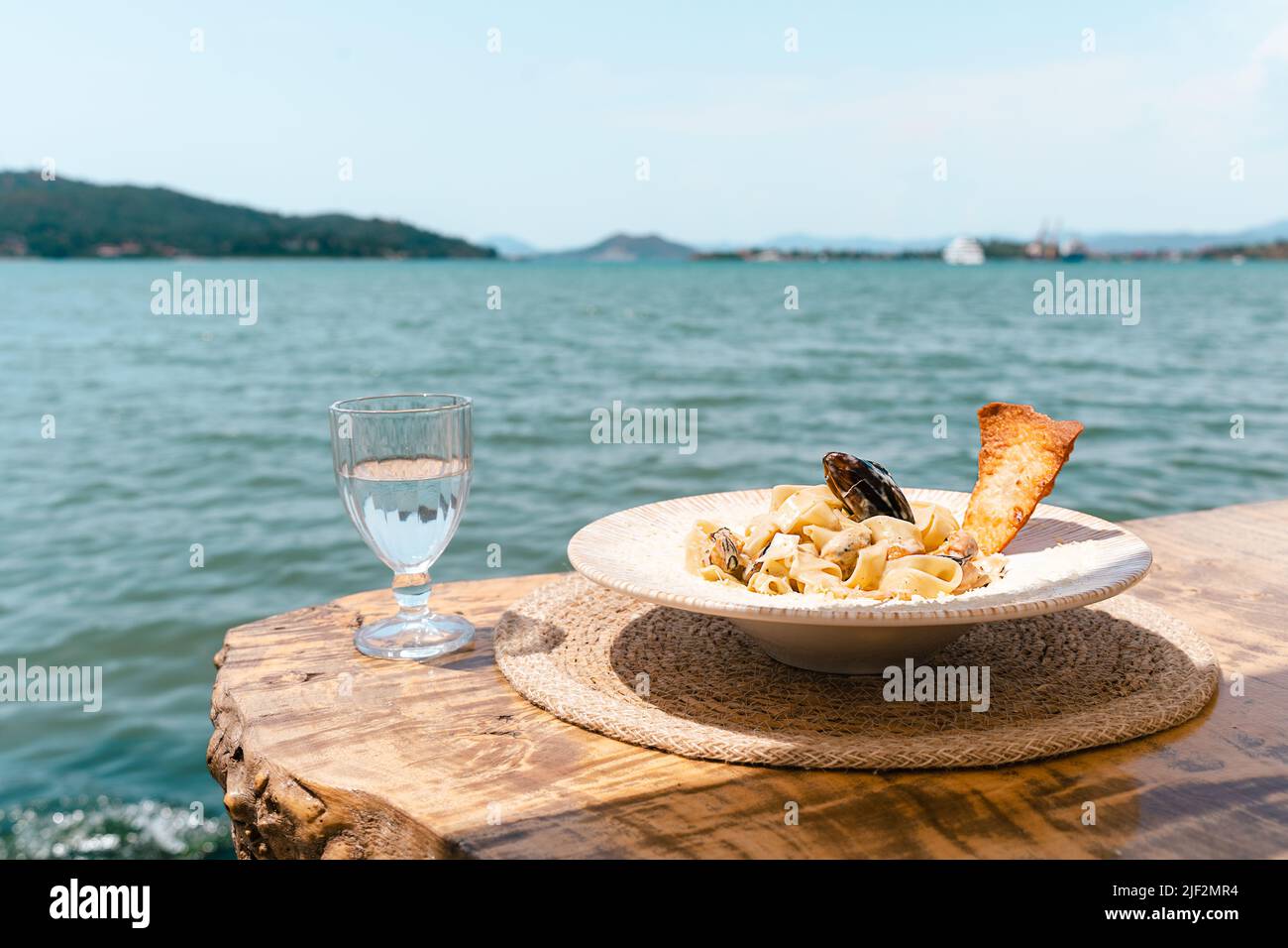 Seafood pasta spaghetti and a glass of water standing on a wooden table in outside cafe restaurant with scenery sea in the background. Copy blank Stock Photo