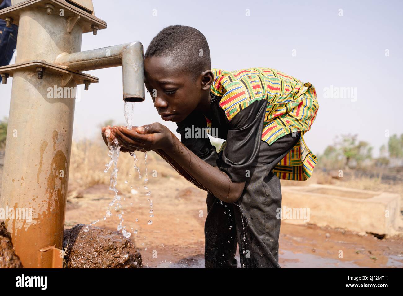 Thirsty young black African boy leans forward to drink fresh and clean water with his bare hands from a tap; water scarcity concept Stock Photo