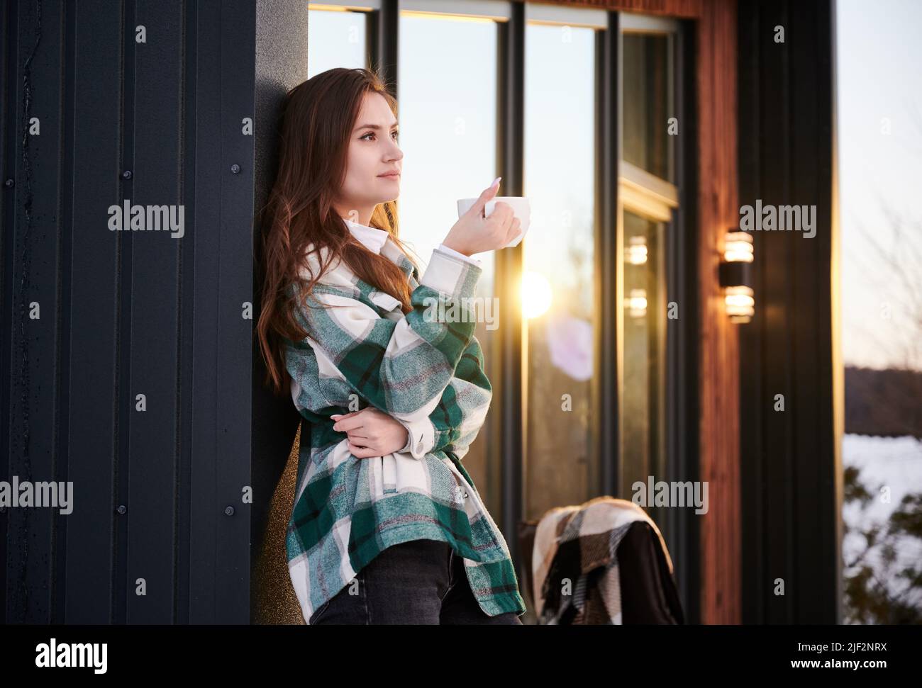 Young woman resting on terrace of modern barn house in the mountains. Happy female tourist holding cup of tea, enjoying winter vacation in new cottage. Stock Photo