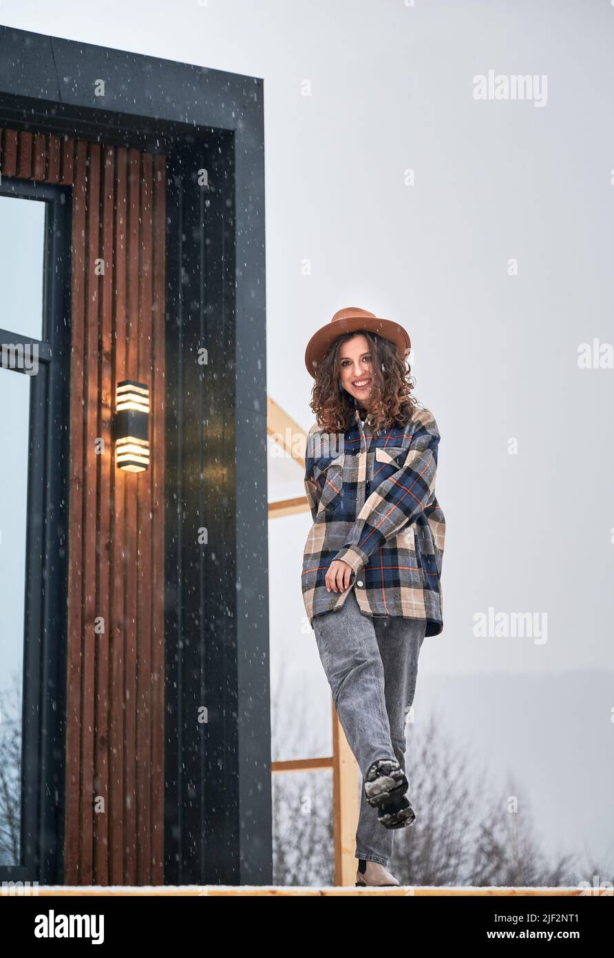 Curly young woman resting on terrace of modern barn house in the mountains. Happy female tourist enjoying winter holidays in new cottage. Stock Photo