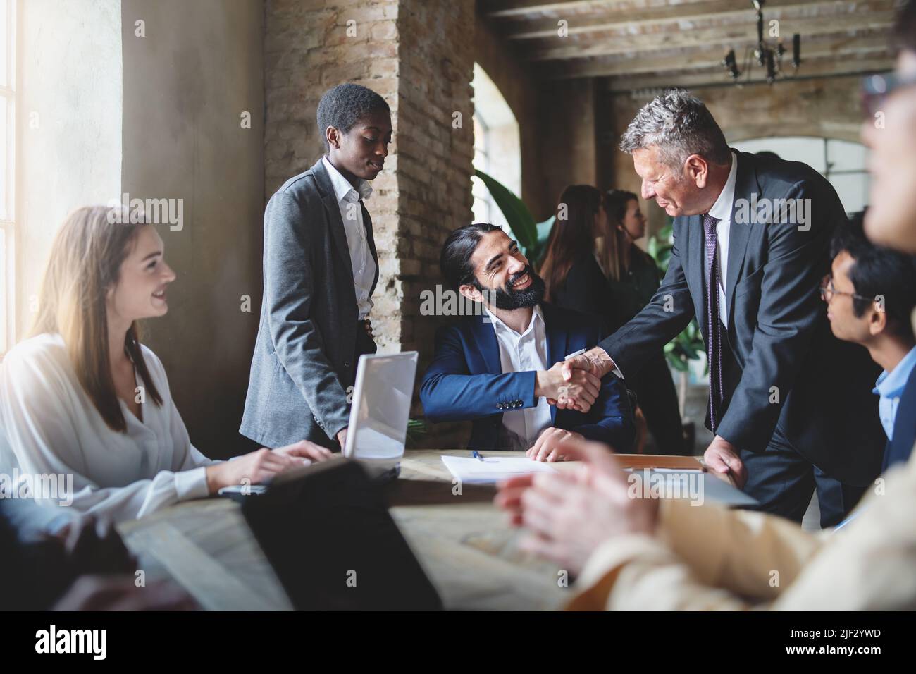 Two businessmen make a deal by shaking hands sitting at a table together with other multiracial employees - business lifestyle and agreements concept Stock Photo