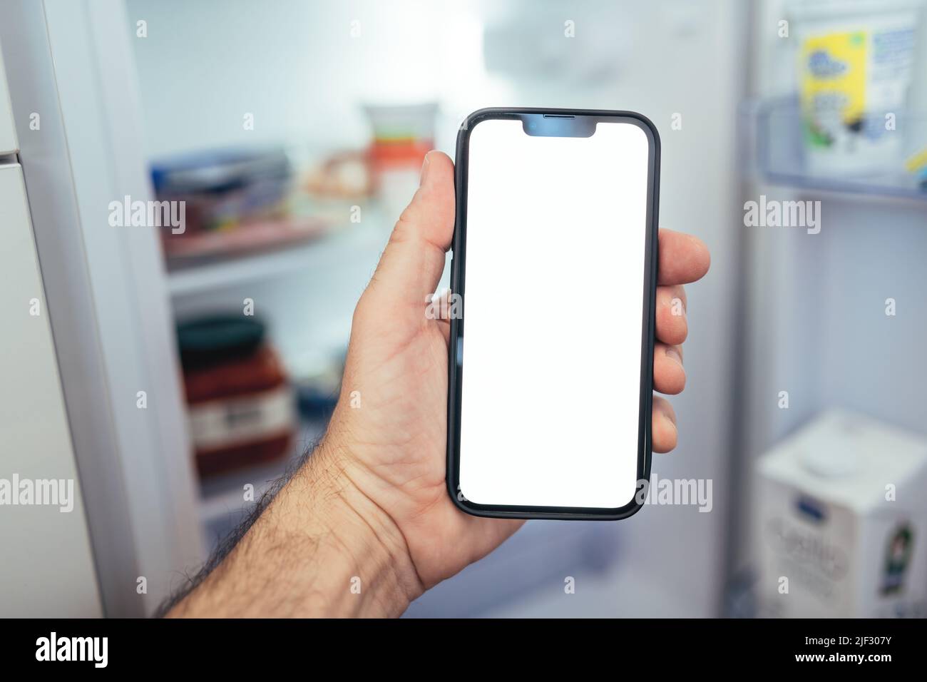 Internet of things smartphone mockup concept, man holding smart mobile phone with blank screen in front of refrigerator and checking on groceries supp Stock Photo