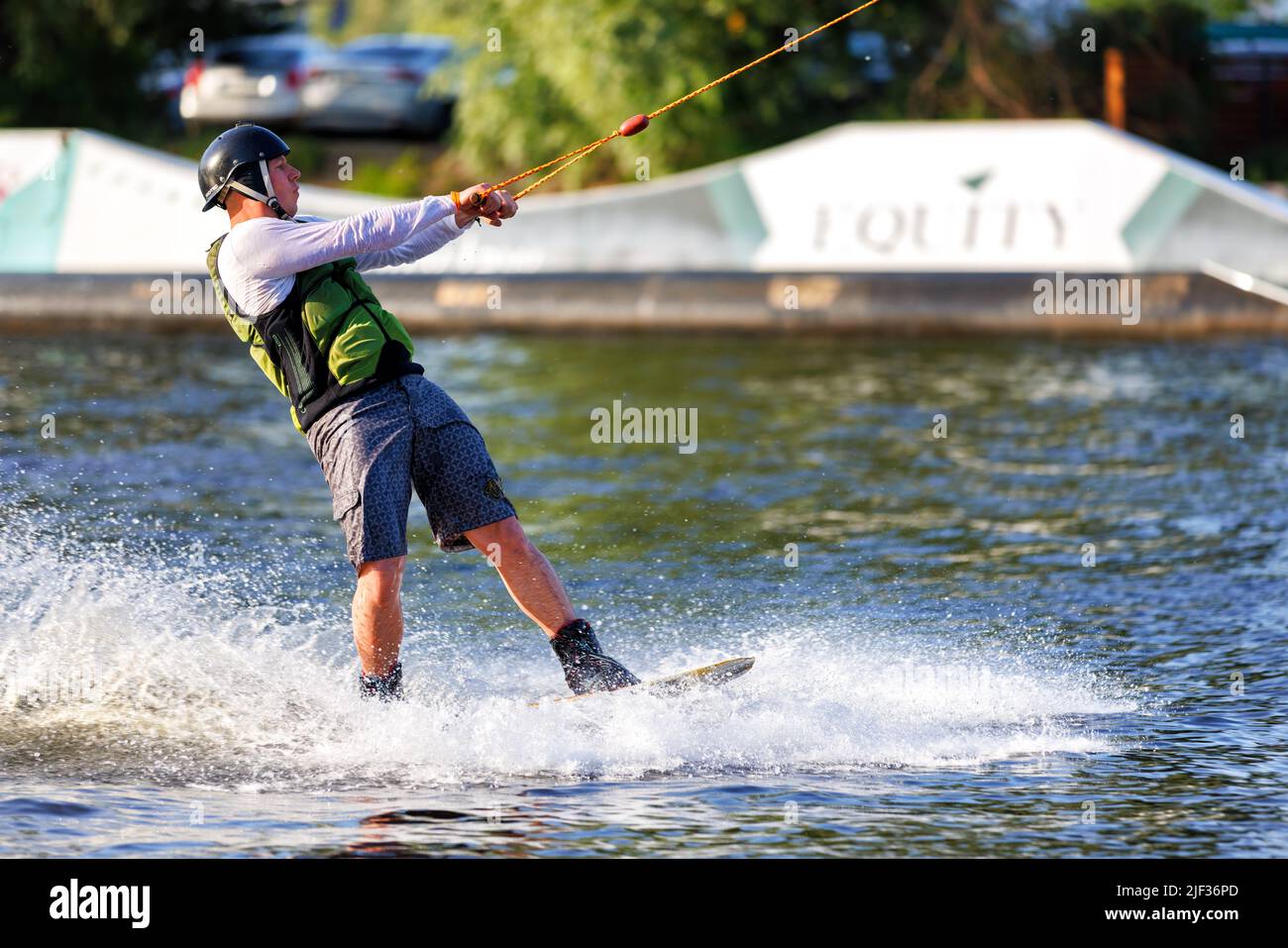 A wakeboarder in a black protective helmet and a green vest rides dynamically on a water board in a summer river bay. 06.19.1922. Kyiv. Ukraine. Stock Photo