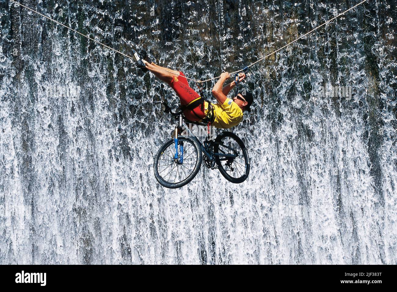 mountainbiker with his bicycle crossing a canyon hanging at a climbing rope, waterfall in the background Stock Photo