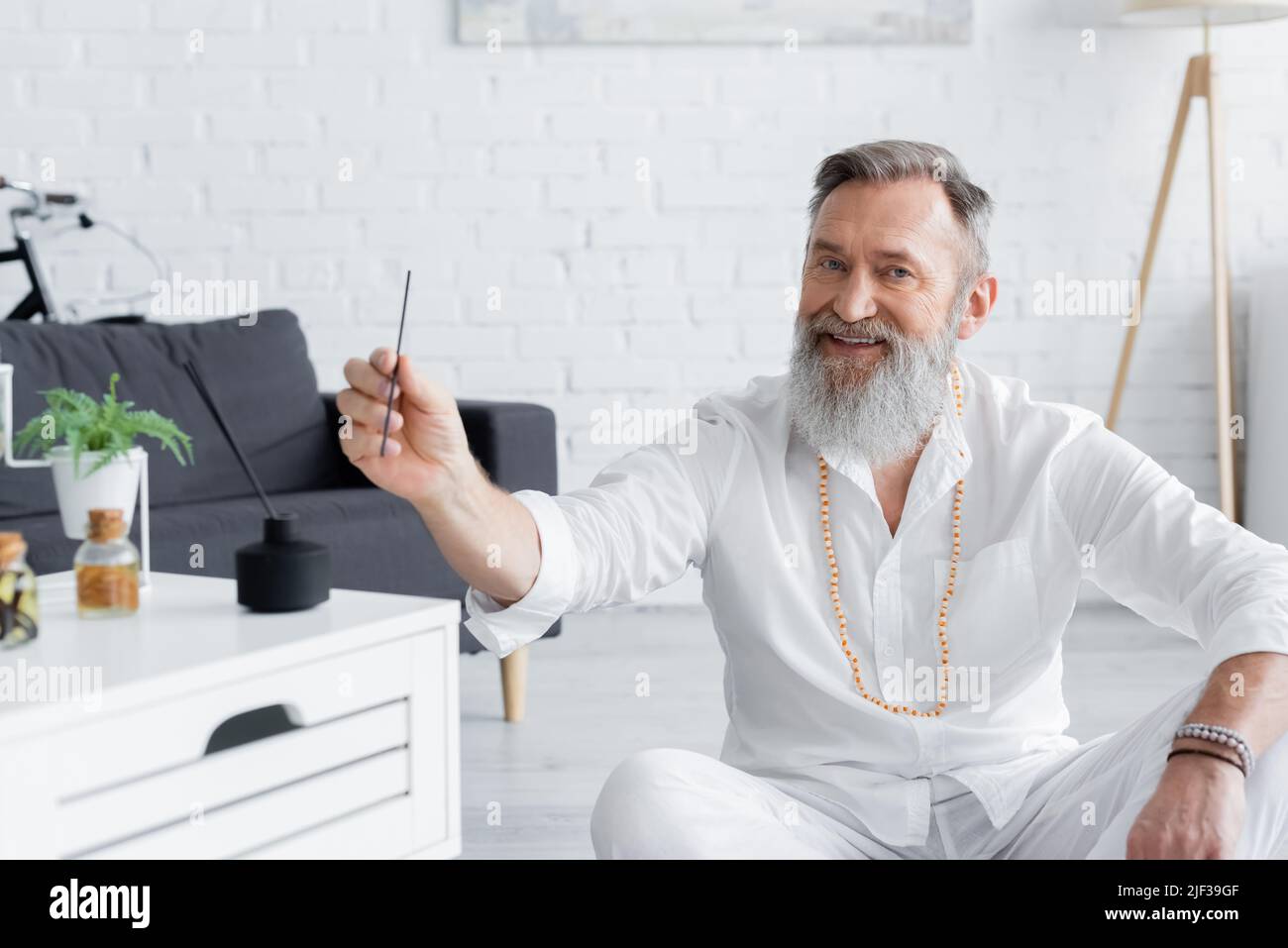 happy ayurveda guru holding aroma stick near blurred nightstand with flavored oils Stock Photo
