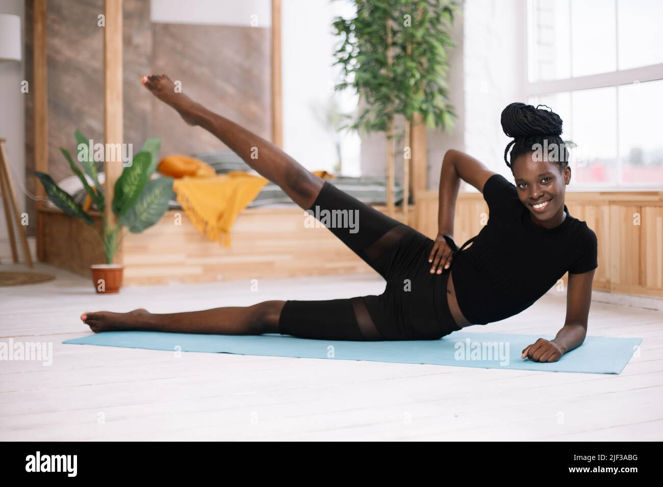 Beautiful athletic afro american woman lying on side and raising leg up selective focus, training on gym mat in decorated photo studio. Sports course Stock Photo