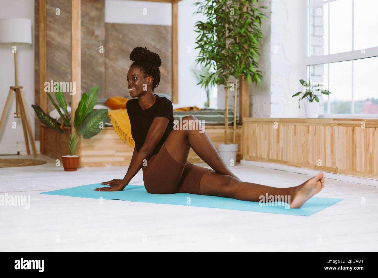 Smiling fit afro american woman turning body sitting on floor with bent leg, training on gymnastic mat in decorated studio. Muscle stretching Stock Photo