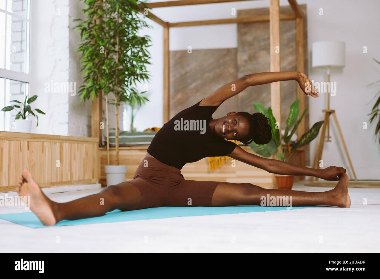 Beautiful and athletic afro american woman sitting and leaning sideways to foot, training on gymnastic mat in decorated studio. Muscle stretching Stock Photo