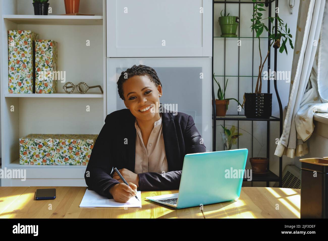 Smiling Young adult entrepreneur freelance black woman small business owner with laptop working in home office looking at camera shot Stock Photo