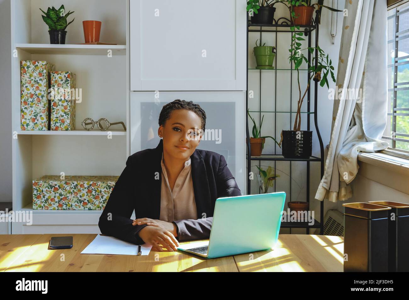 Young adult entrepreneur freelance black woman small business owner with laptop and smartphone working in home office looking at camera shot Stock Photo