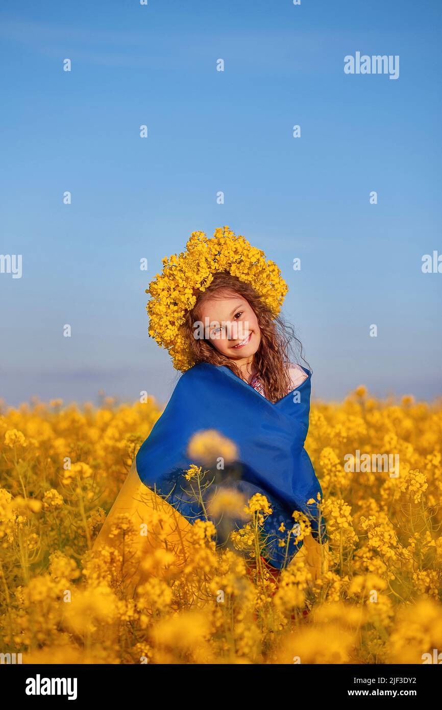 Ukrainian child girl wrapped in blue and yellow Ukrainian flag in yellow wreath in field of yellow flowers against blue sky. Pray for Ukraine. Ukraine's Independence Flag Day. Symbols of Ukraine. Stock Photo