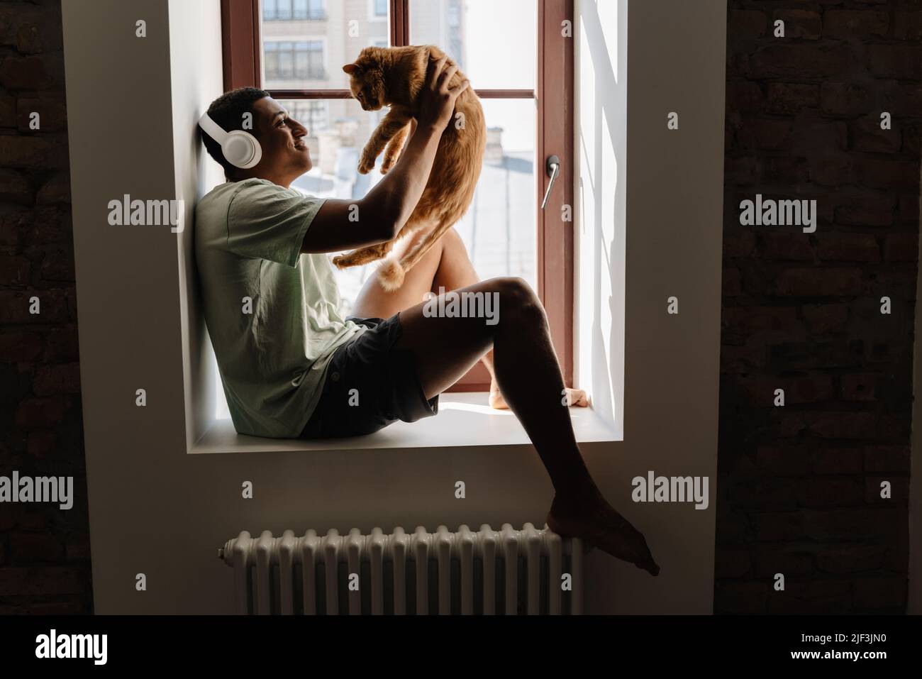 Black man in headphones sitting with his cat on windowsill in hotel Stock Photo