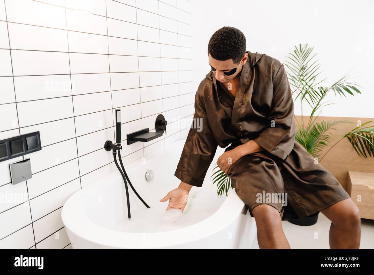 Black man dressed in silk robe using eye patches while sitting on bath ...