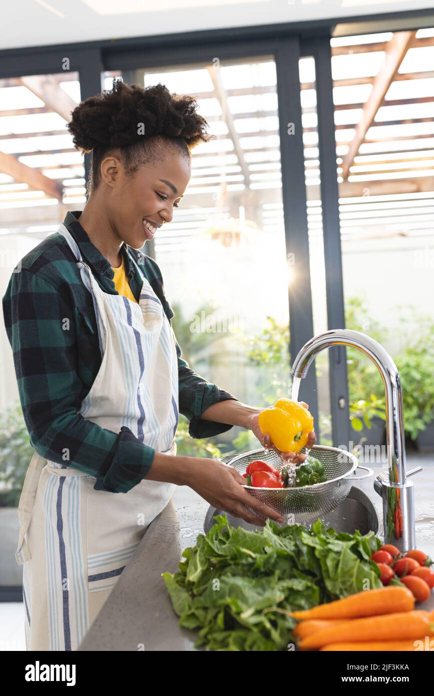 Smiling african american mid adult woman wearing apron cleaning vegetables in kitchen sink Stock Photo
