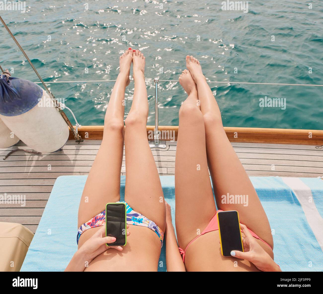 Two friends sunbathing on boat during holiday cruise together using cellphones to send text messages. Two women lying on boat using smartphones and Stock Photo