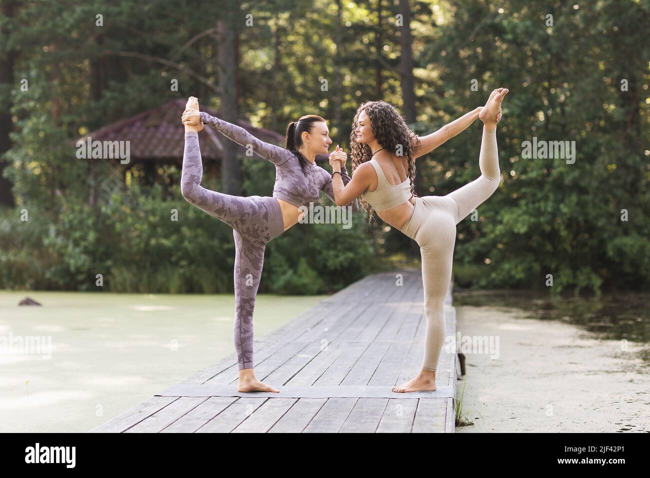 Two women holding hands perform the Natarajasana exercise, the pose of the king of dancers, they train in the morning on a wooden bridge in a summer p Stock Photo