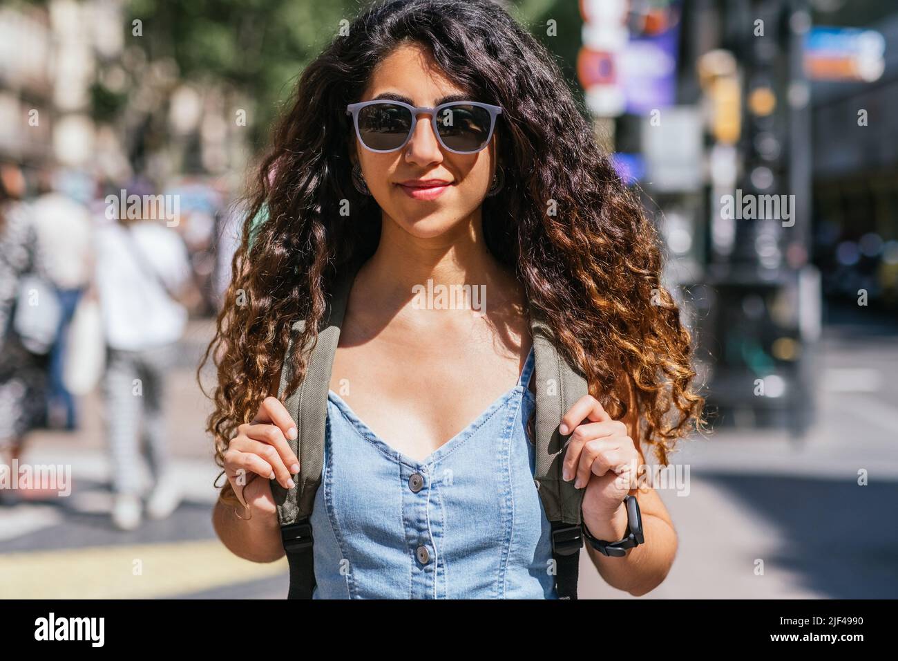 portrait of a beautiful dark haired young woman wearing denim dress. She wears sunglasses and a backpack and looks to camera positively Stock Photo