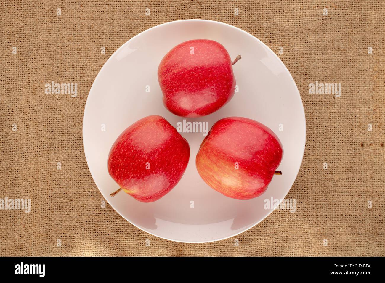 Four juicy red apples with a white ceramic plate on a jute cloth, close