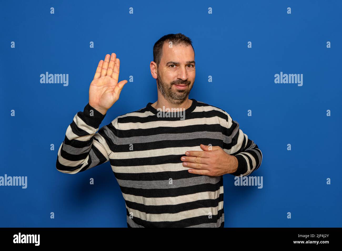 Hispanic man with beard and striped sweater standing over isolated blue background Swearing with hand on chest and open palm, making a loyalty promise Stock Photo