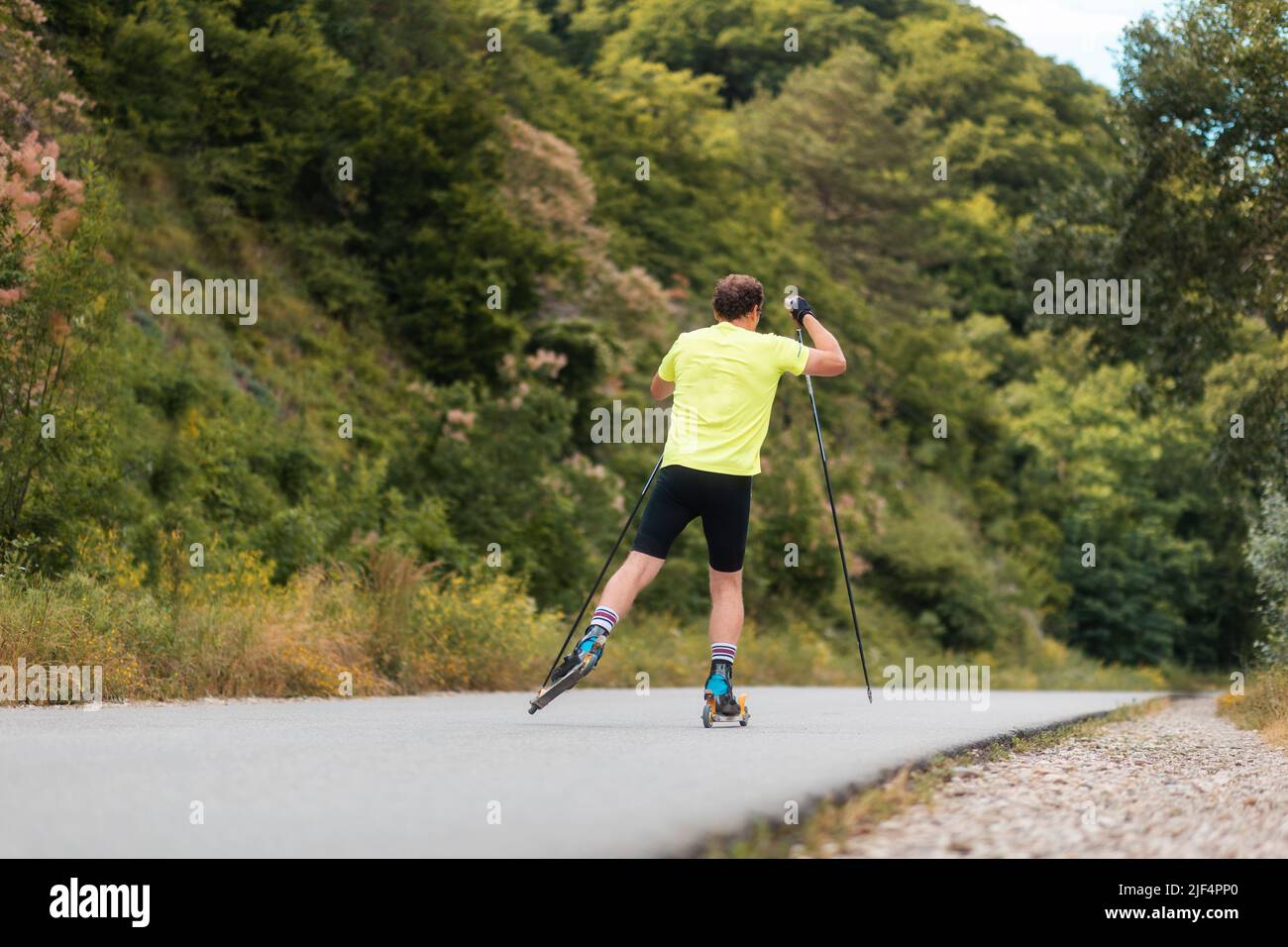 Biathlon workout. Athletic man training on the roller ski at country road, back view. Concept of competition and summer activity. Stock Photo