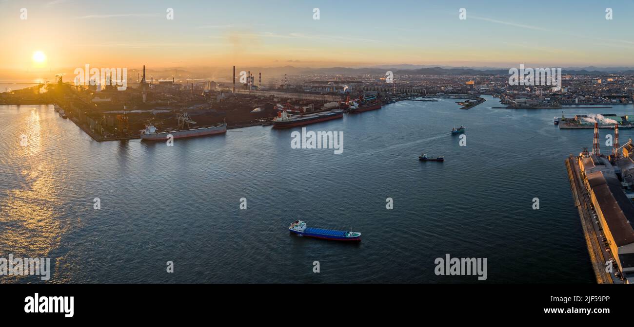 Panorama: Aerial view of ships docked and anchored at industrial harbor as sun sets Stock Photo