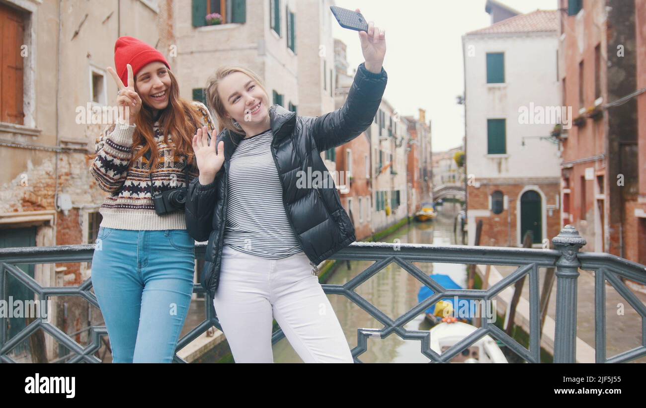 Two young women in warm clothes standing on a background of a water canal and taking a selfie - Venice, Italy. Mid shot Stock Photo