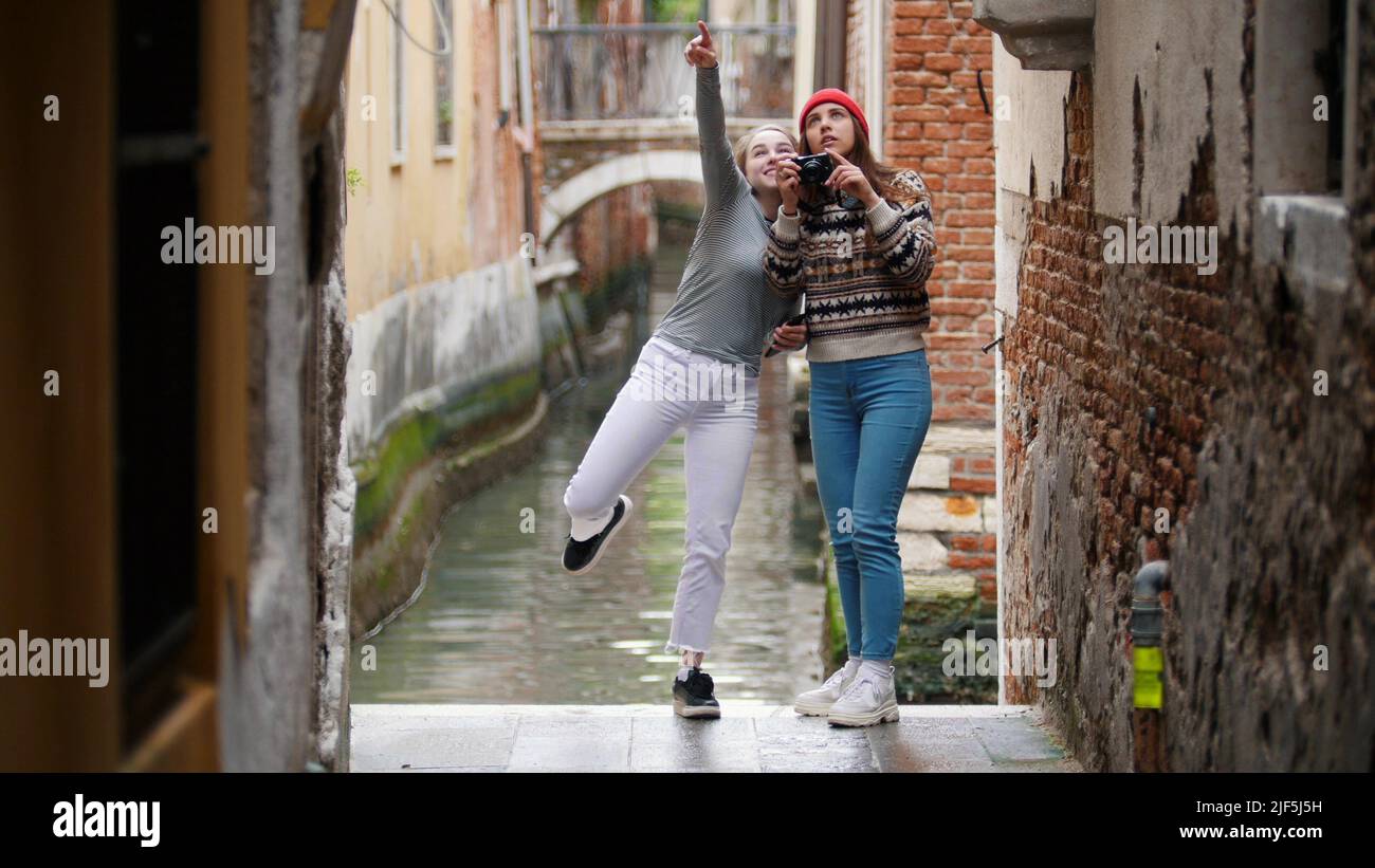 Two young women travelers in warm clothes standing on a background of a water canal and taking photos - Venice, Italy. Mid shot Stock Photo