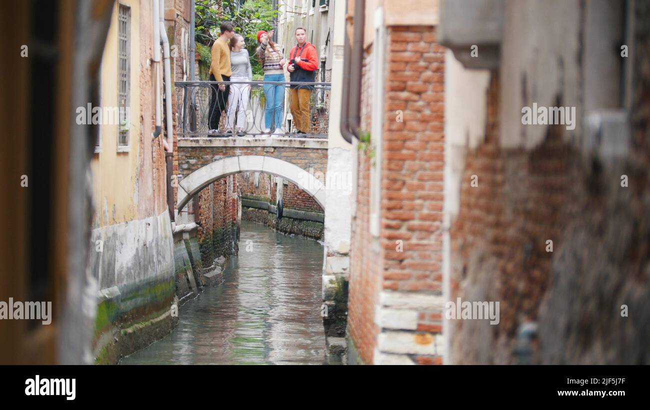 Four young friends standing on the bridge between two buildings above the water channel - Venice, Italy. Mid shot Stock Photo
