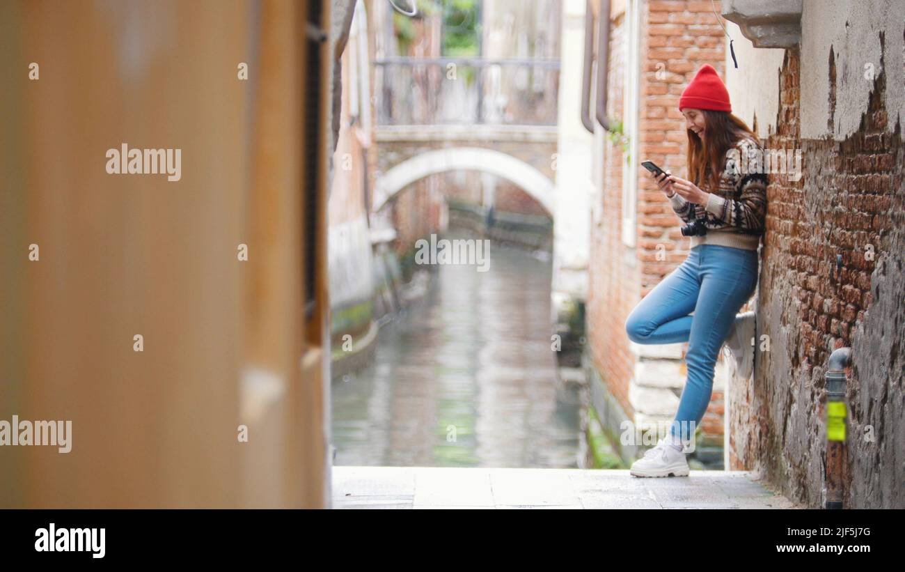 A young woman standing near the water channel and searching for something in her phone - Venice, Italy. Mid shot Stock Photo