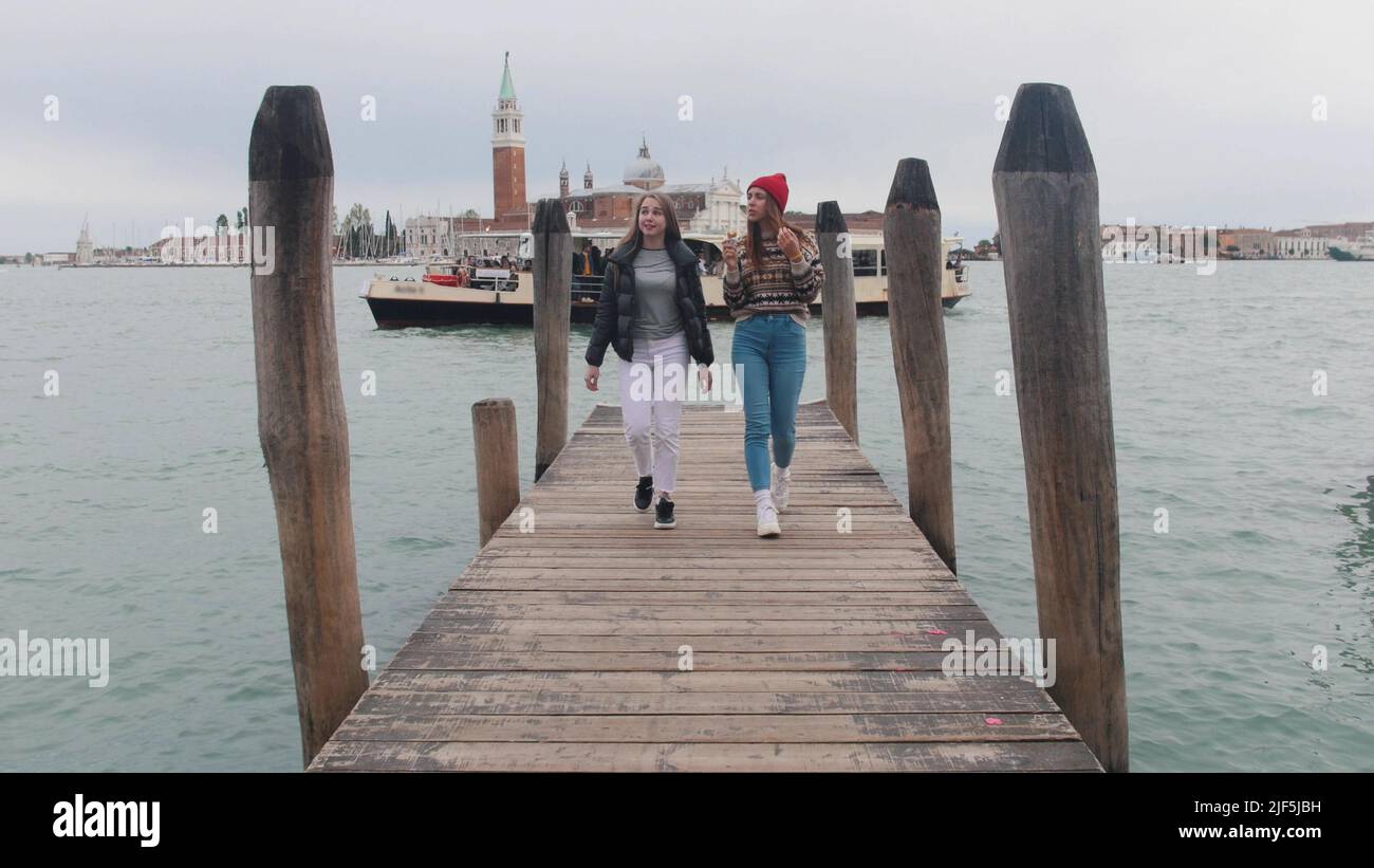 Two young women walking on the pier. One of them eating icecream. Mid shot Stock Photo