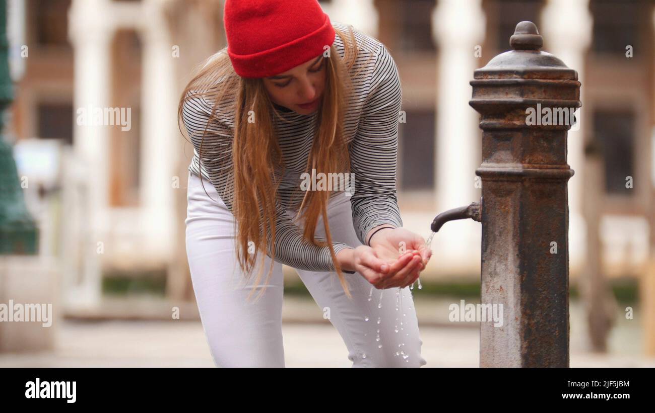 A young woman drinks water from a street fountain - filling her hands with water. Mid shot Stock Photo