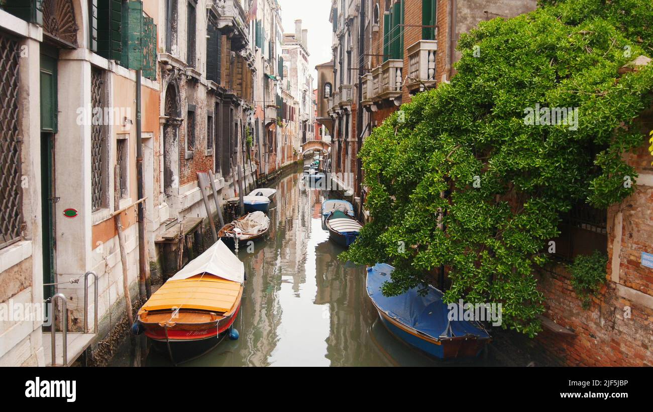 Narrow streets of Venice with canal filled with water - moored boats. Mid shot Stock Photo