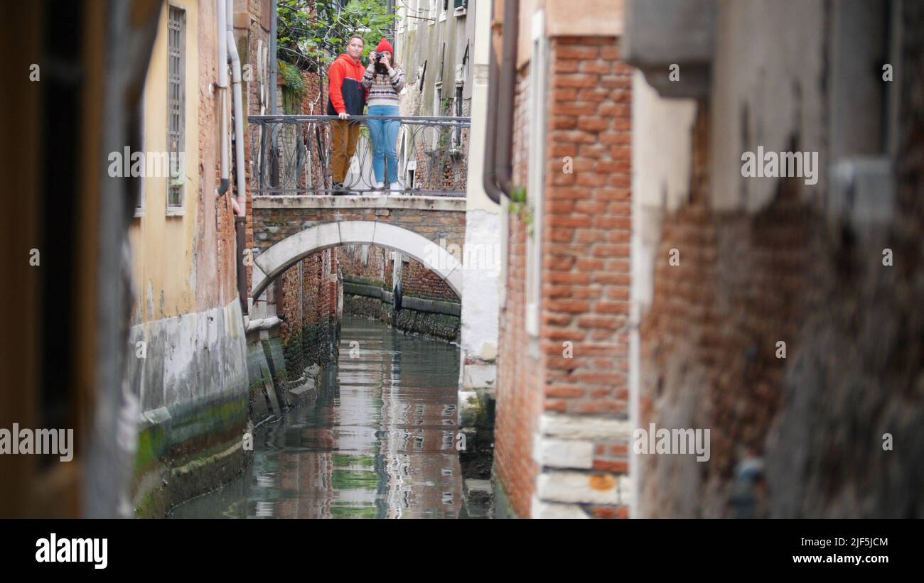 A young couple standing on the bridge between two buildings and taking photo - Venice, Italy. Mid shot Stock Photo