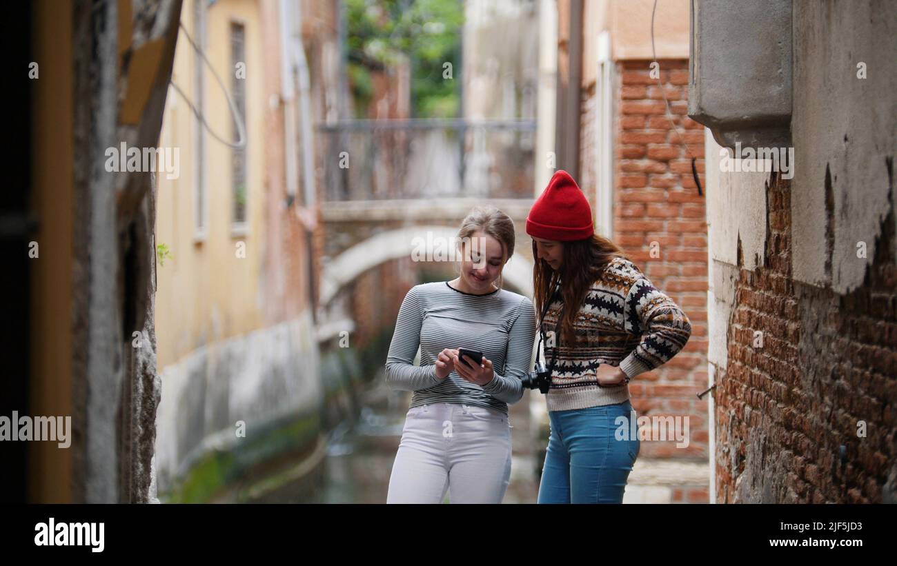 Two young women standing near the water channel and looking at the phone screen - Venice, Italy. Mid shot Stock Photo