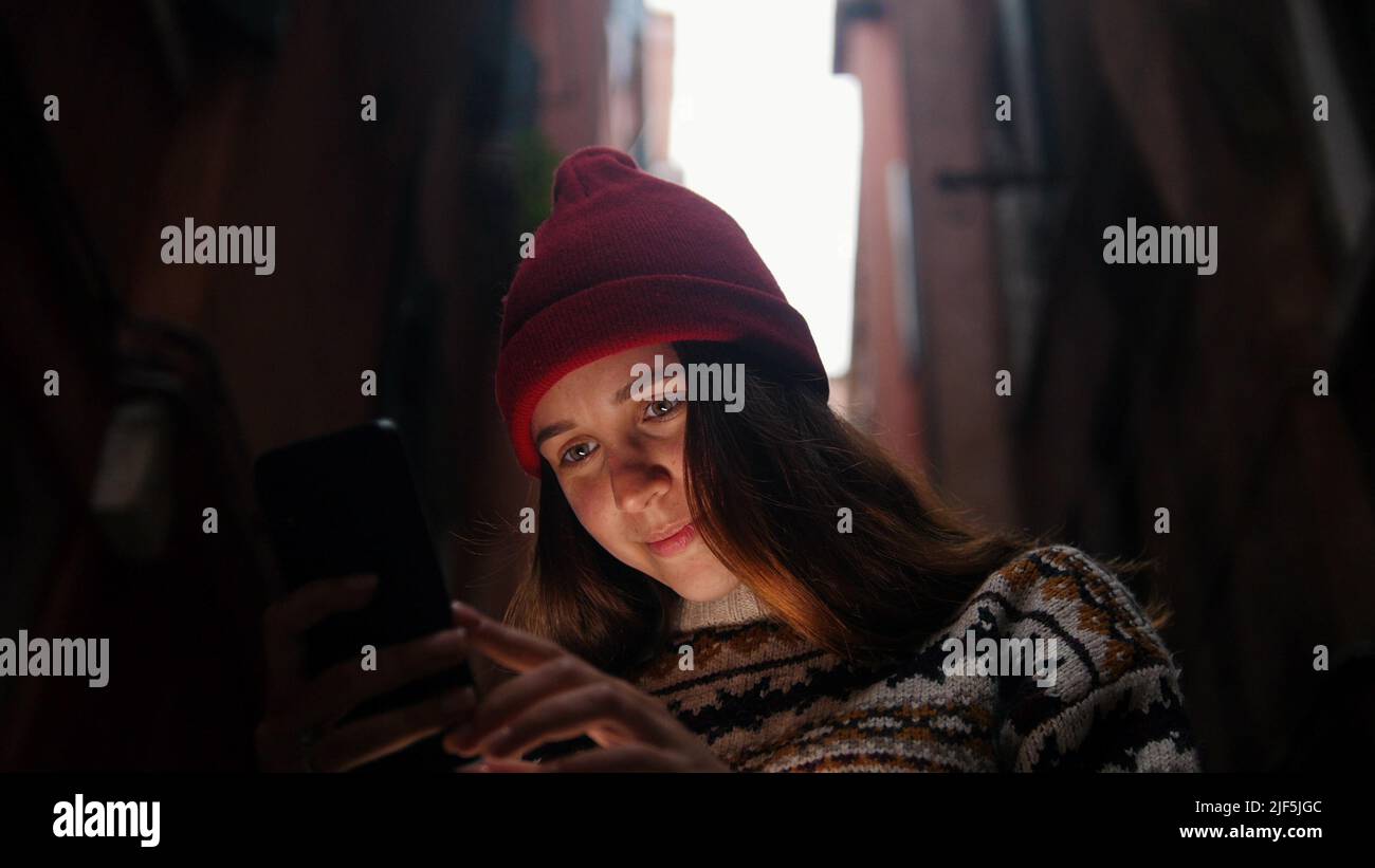 Woman in a dark alley between buildings. Looks into her phone - Mid shot Stock Photo