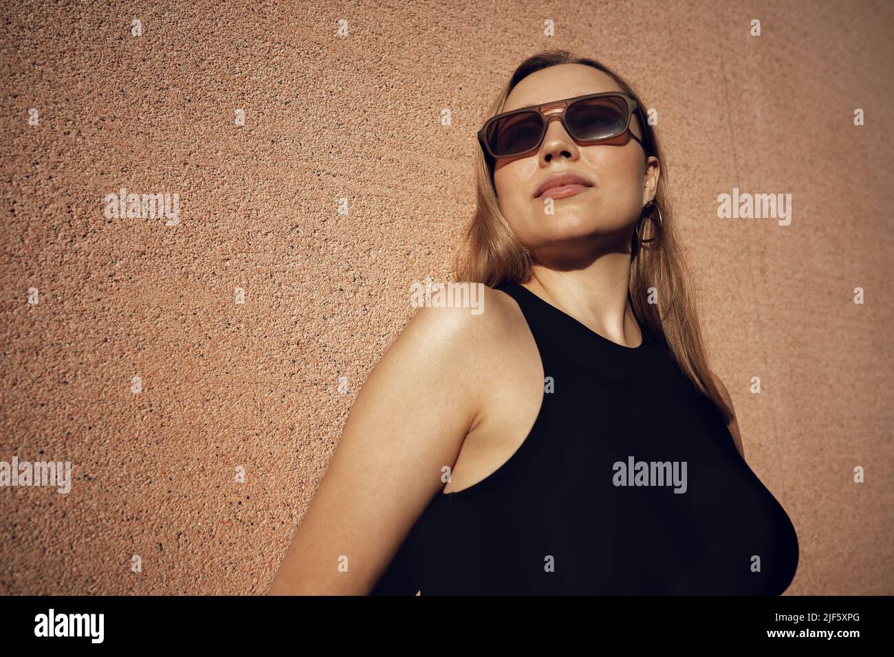 The blonde model wearing sunglasses stands against the beige wall Stock Photo