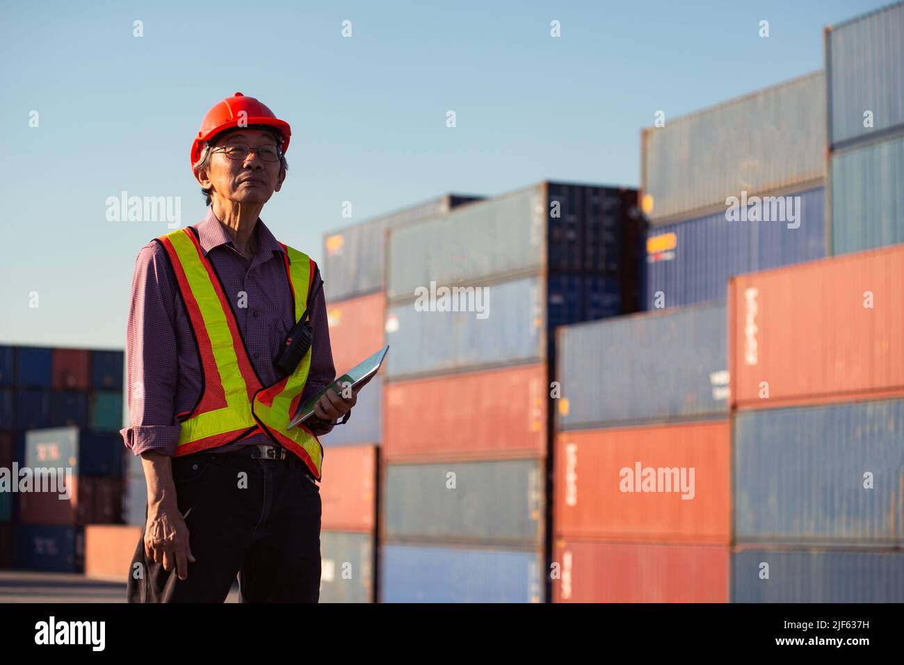 A senior elderly Asian worker engineer wearing safety vest and helmet standing and holding digital tablet at shipping cargo containers yard. Stock Photo