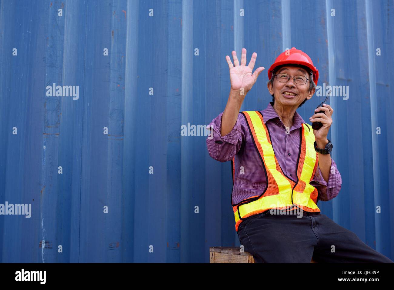 A senior elderly Asian worker engineer wearing safety vest and helmet standing and holding radio walkies talkie at logistic shipping cargo containers Stock Photo