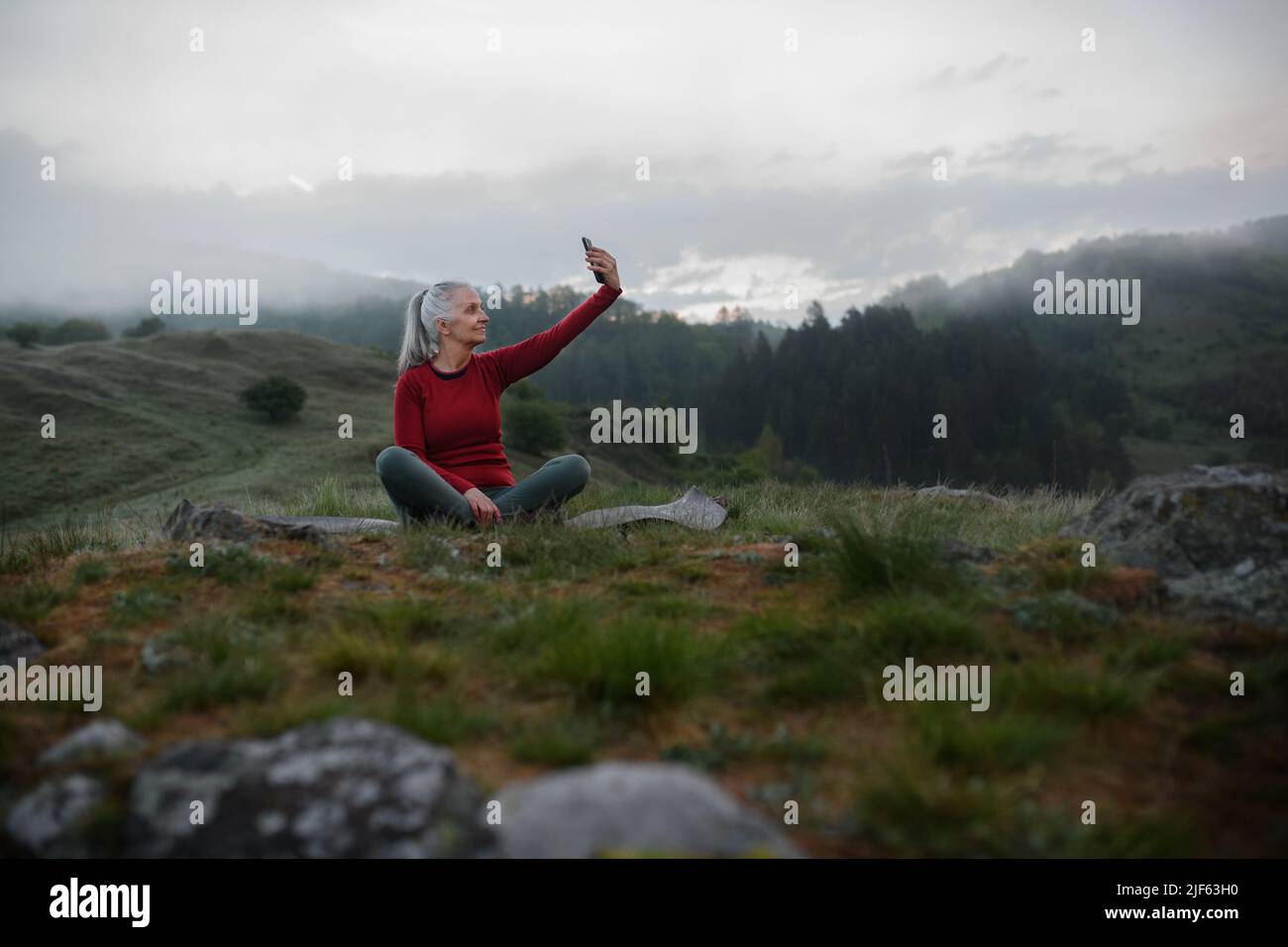 Senior woman taking selfie when doing breathing exercise in nature on early morning with fog and mountains in background. Stock Photo